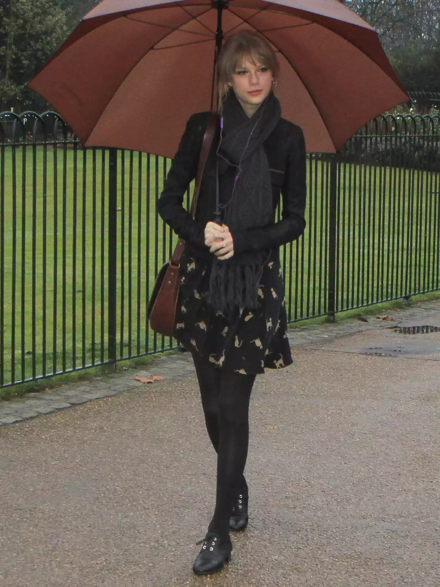 Taylor Swift wearing black tights, black flats, a black mini dress, scarf, and brown leather bag while walking with a brown umbrella in London's Hyde Park