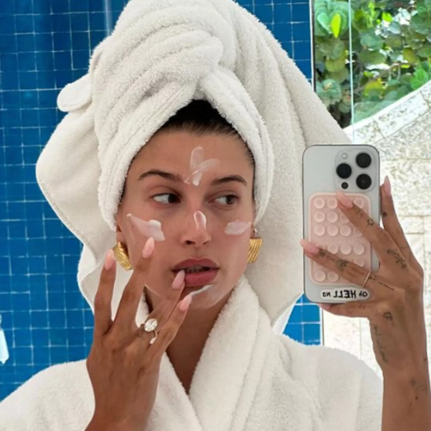 Hailey Bieber taking a selfie in a robe with her hair up in a towel, applying skincare with her light pink lip gloss nails.