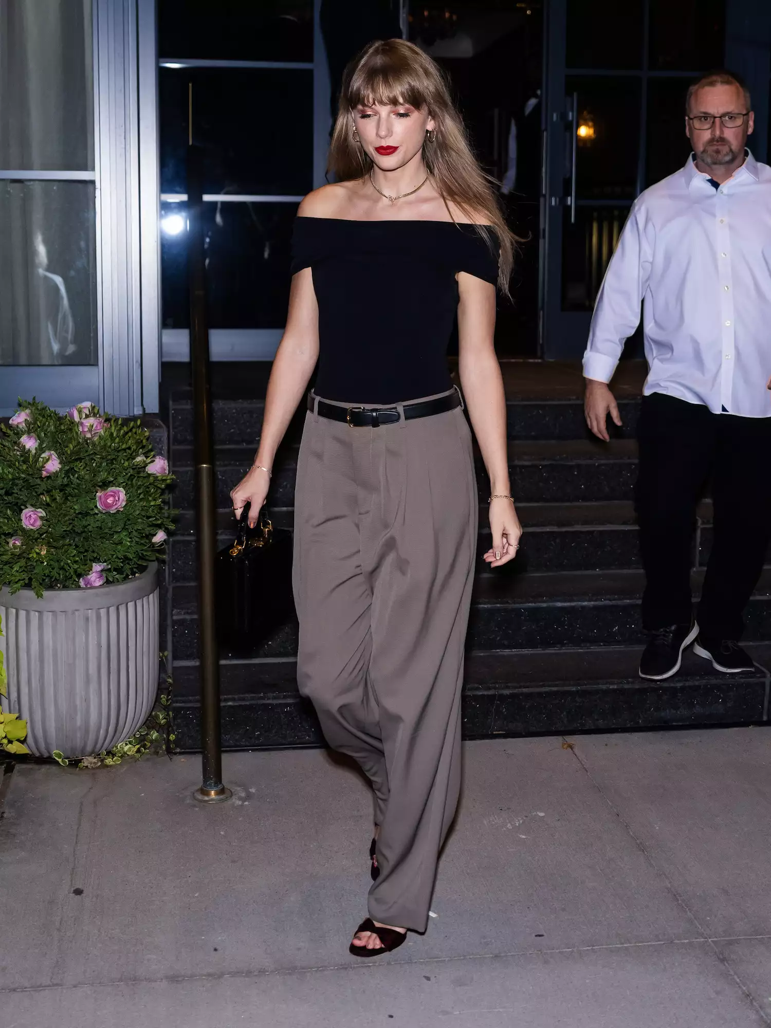 Taylor Swift wears a black boat-neck top, gray trousers, a black top handle bag, open toe heels, and a chain necklace